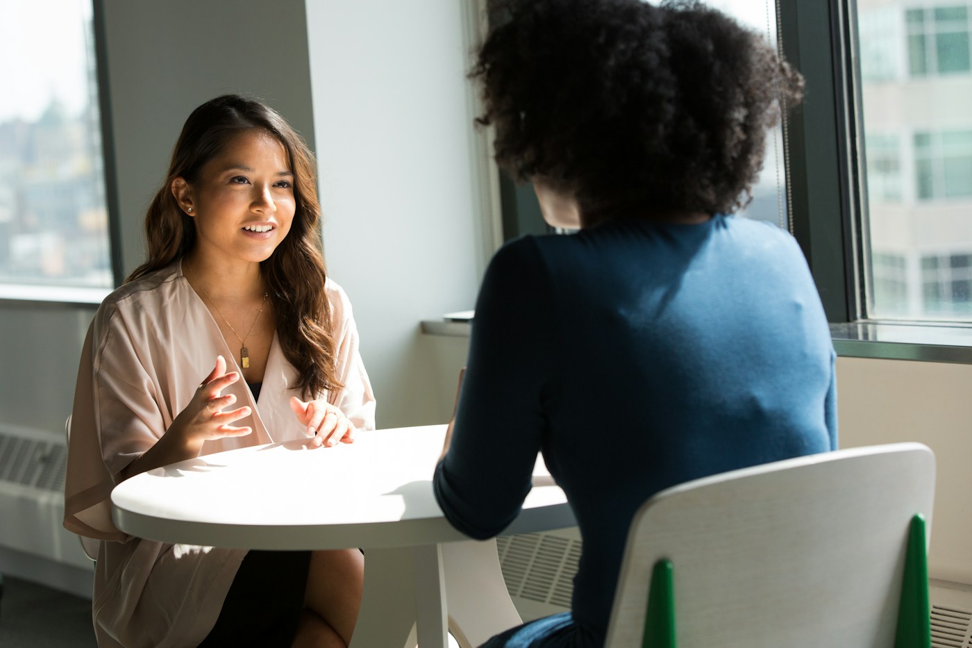 Two adult women, including a Black professional, sharing a relaxed conversation at a sunlit table.
